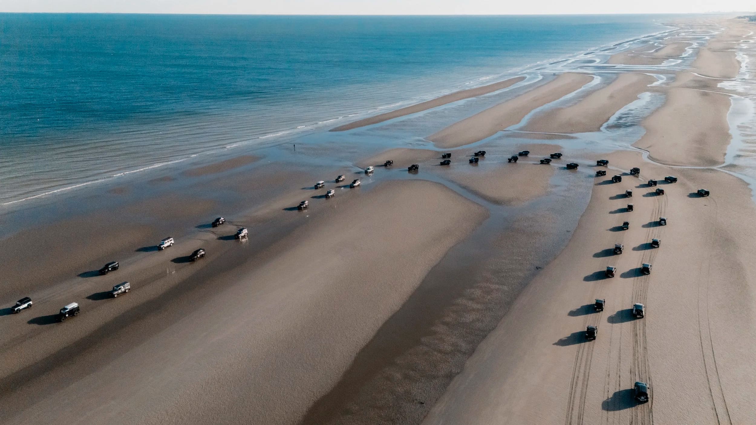 Aerial view of a convoy of 4x4s driving in lines across a wide beach at low tide.