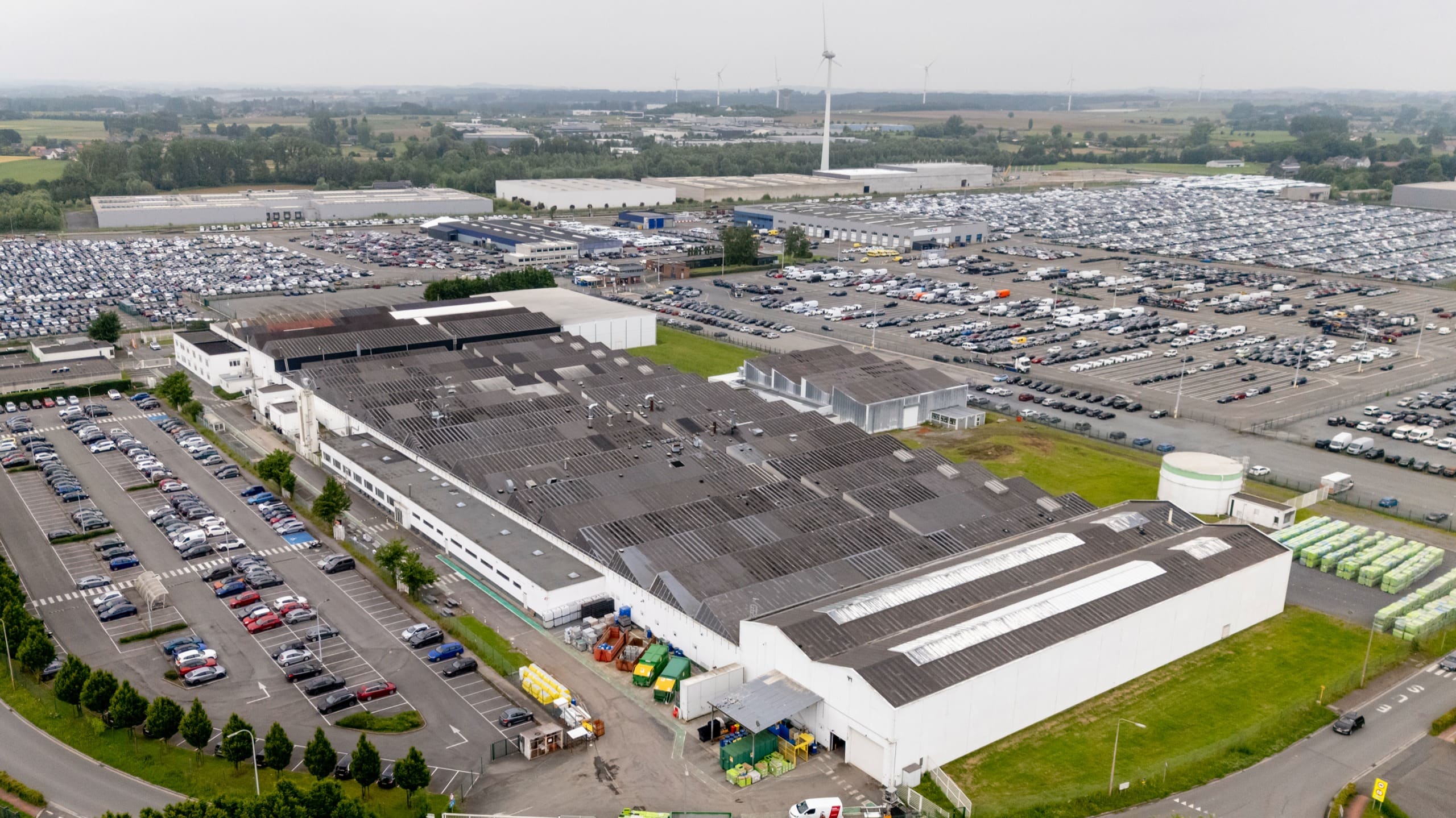 Vue aérienne d’une usine aux longues toitures à sheds, avant les travaux, entourée de vastes parkings.
