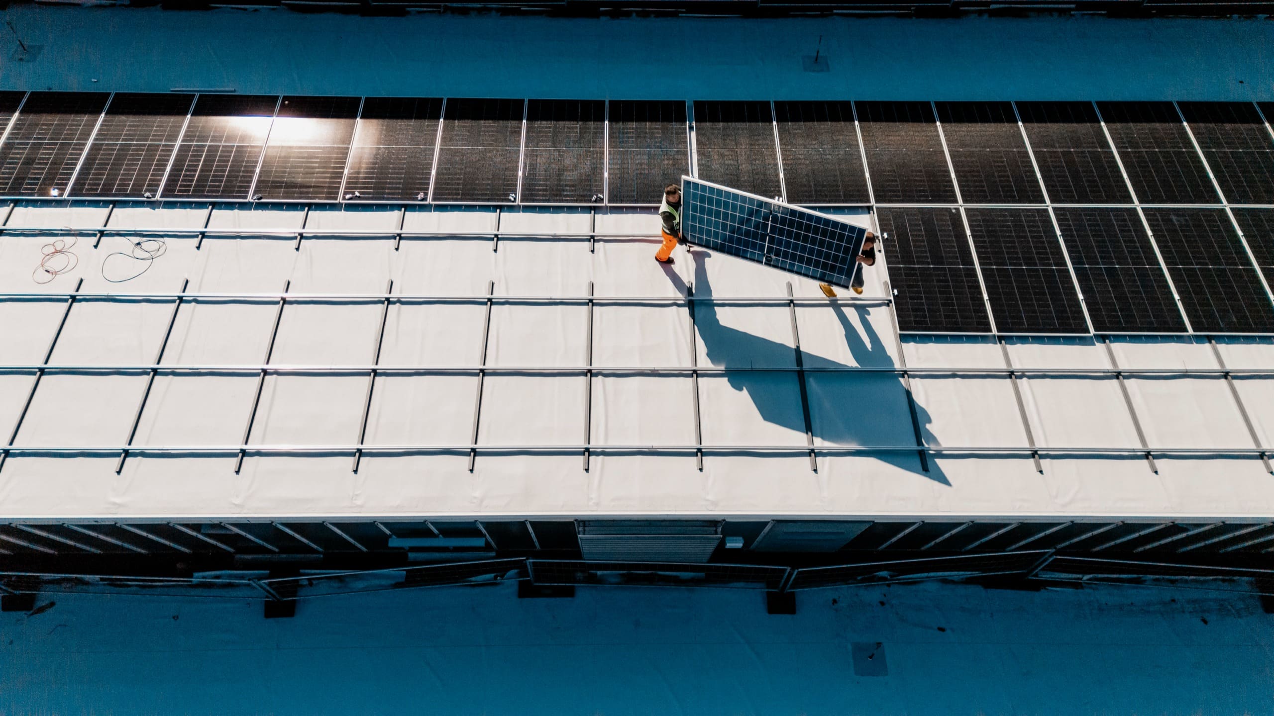 Vue d’en haut : un ouvrier porte un panneau solaire sur une toiture blanche, à côté des rangées déjà posées.