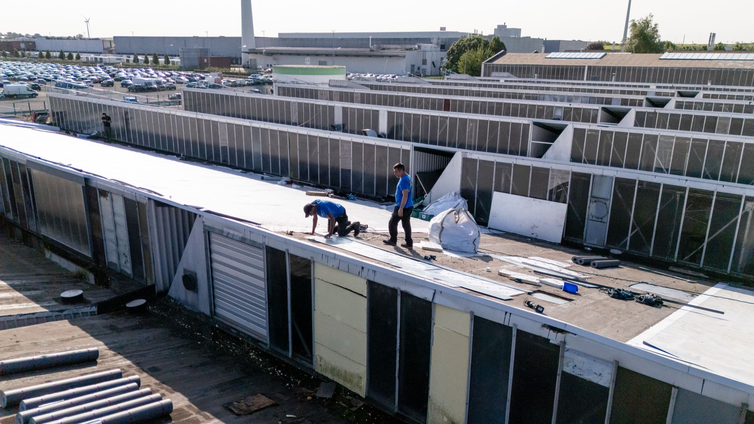 Vue d’en haut : des couvreurs au travail entre les rangées de sheds d’une grande toiture industrielle.