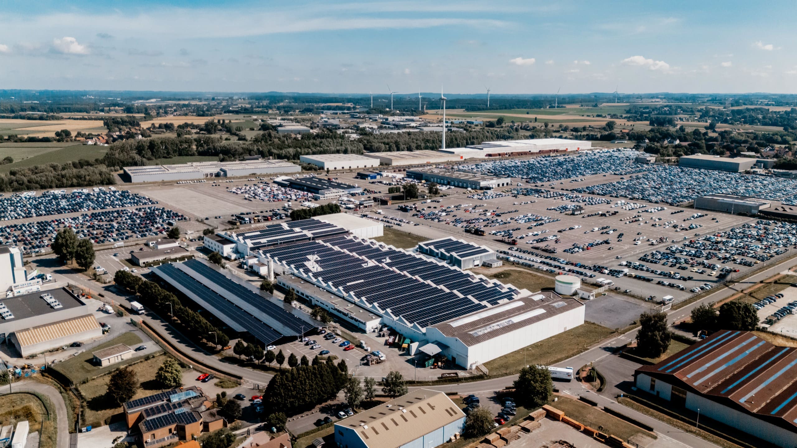 Vue aérienne d’un site industriel aux toitures couvertes de panneaux solaires, entre parkings et campagne.