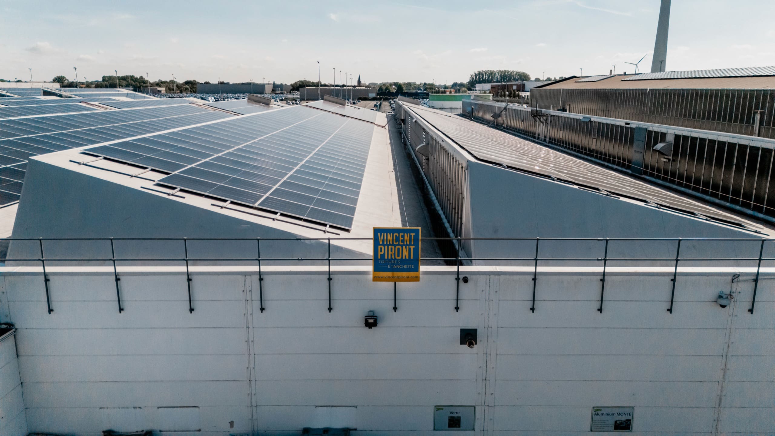 Le haut d’un bâtiment industriel aux toitures solaires, avec l’enseigne de l’entreprise de toiture.