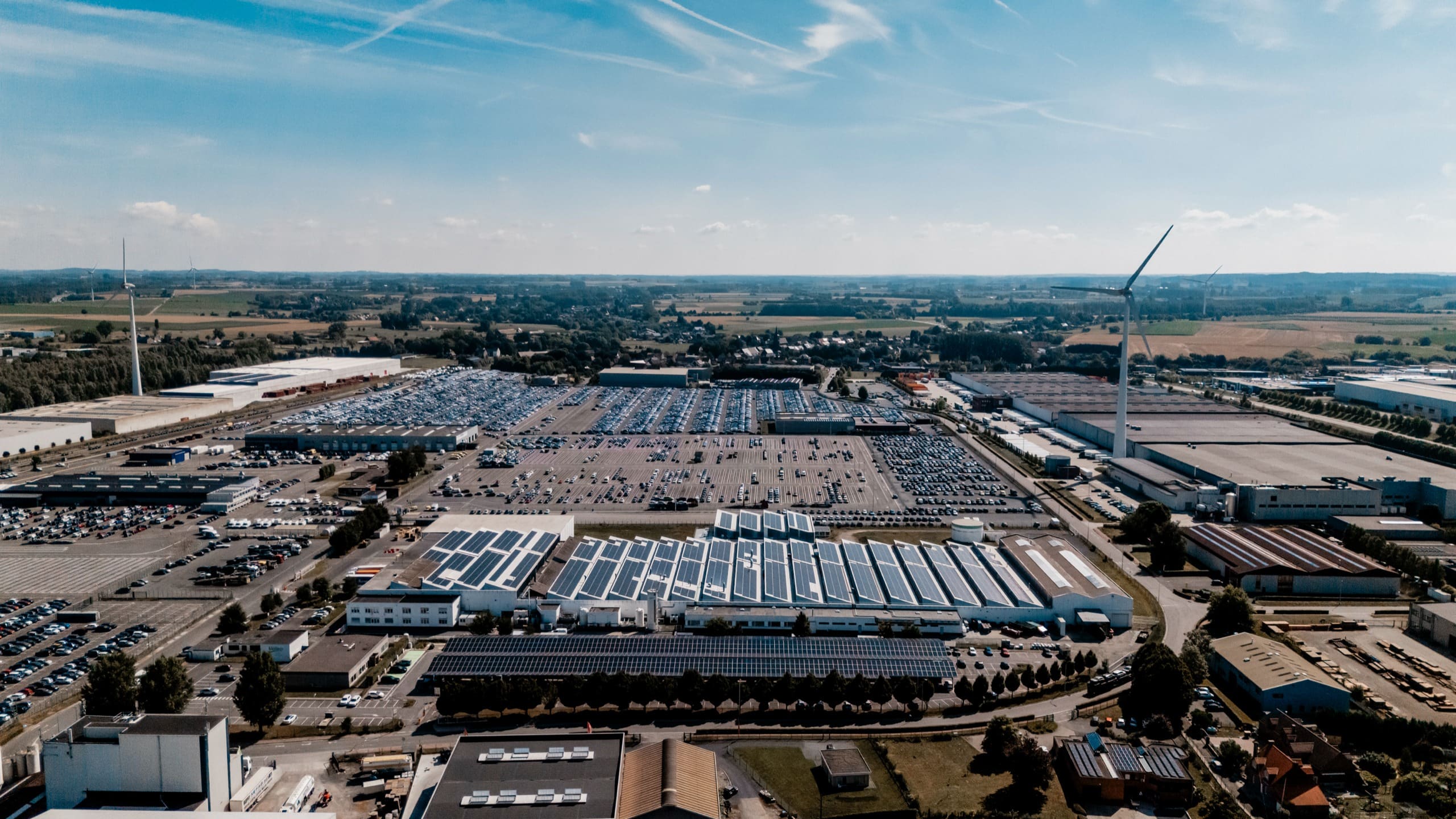 Vue aérienne lointaine du site industriel et de ses toitures solaires, des éoliennes à l’horizon.