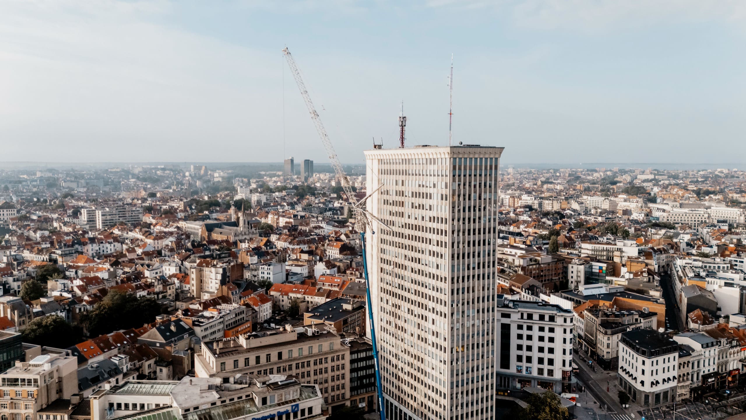 Une tour de bureaux blanche au-dessus des toits de Bruxelles, une grue dressée à son pied, au petit matin.