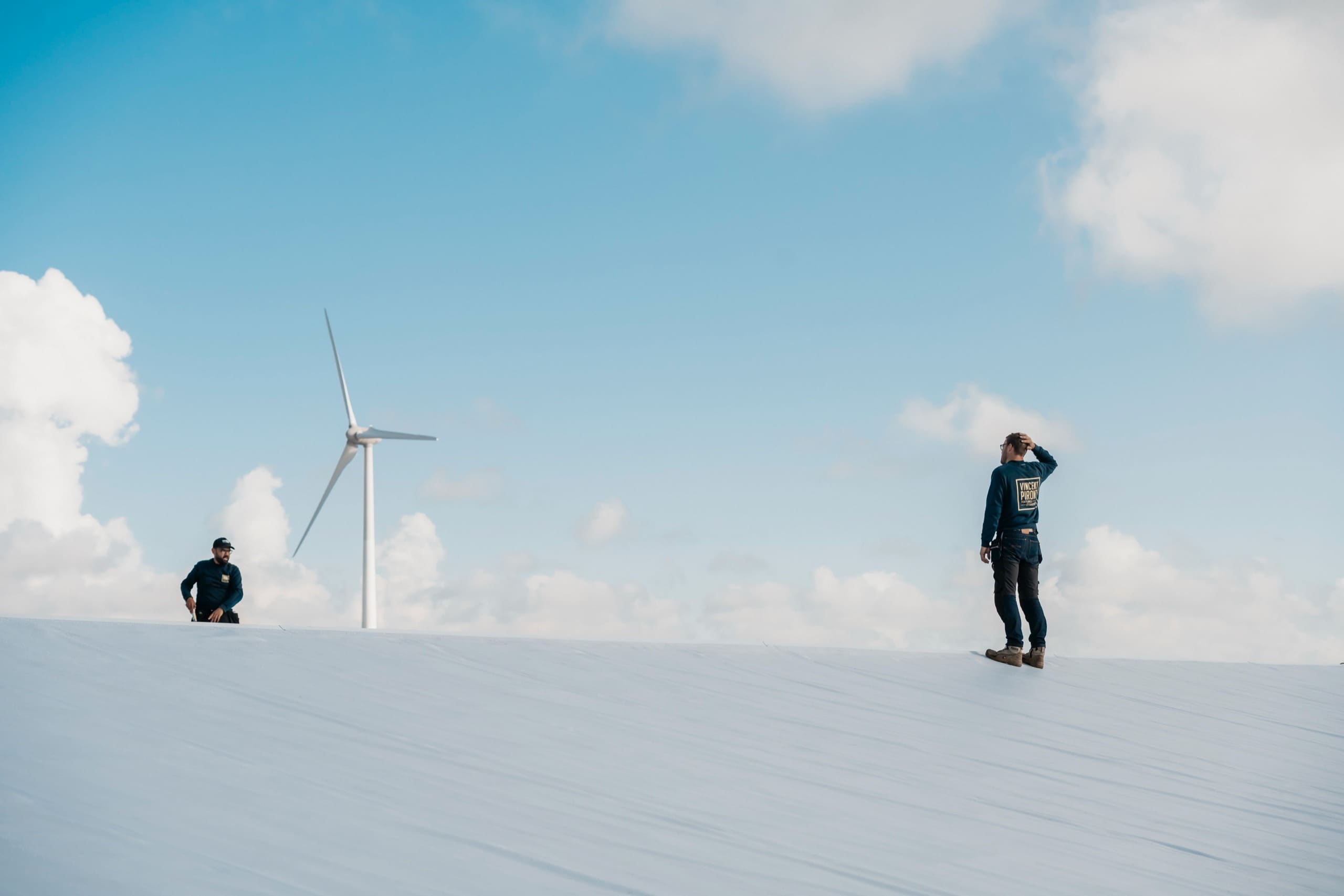 Deux couvreurs sur une toiture blanche, une éolienne et des nuages en arrière-plan.