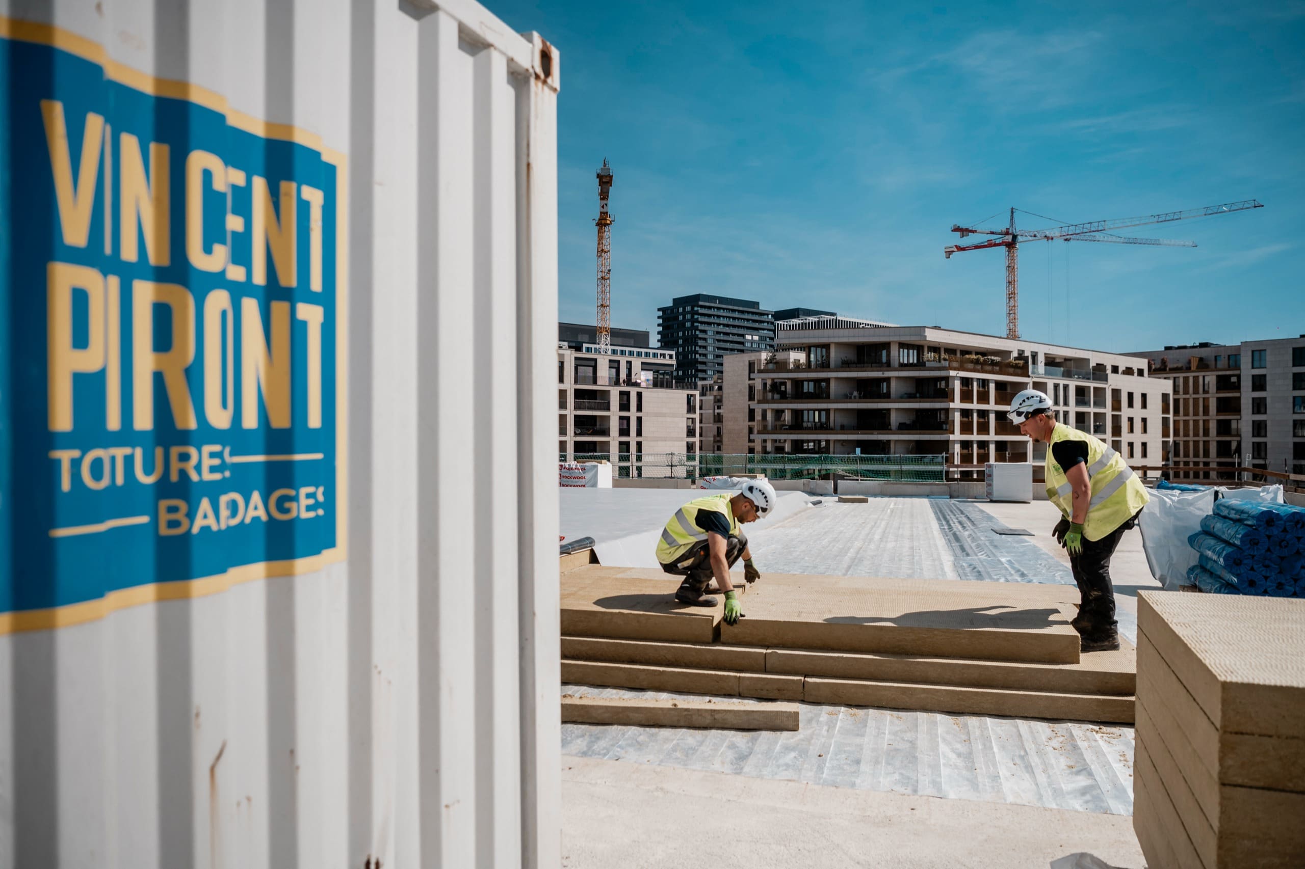 Deux couvreurs posent des panneaux d’isolation sur un toit, près d’un conteneur au logo de l’entreprise de toiture, des grues dans le ciel bleu.