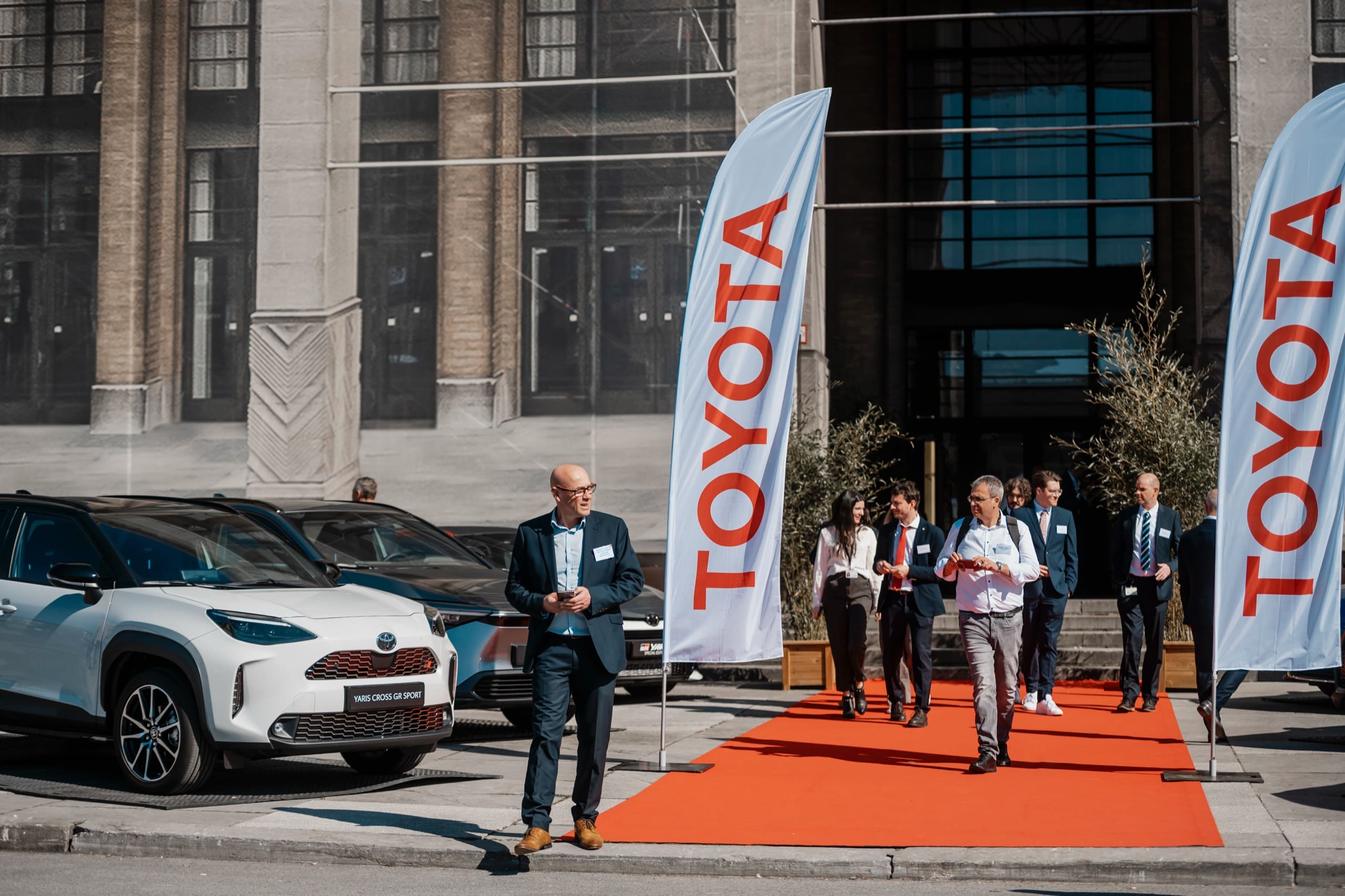 Les invités remontent le tapis rouge entre les drapeaux, une voiture exposée devant l’entrée.