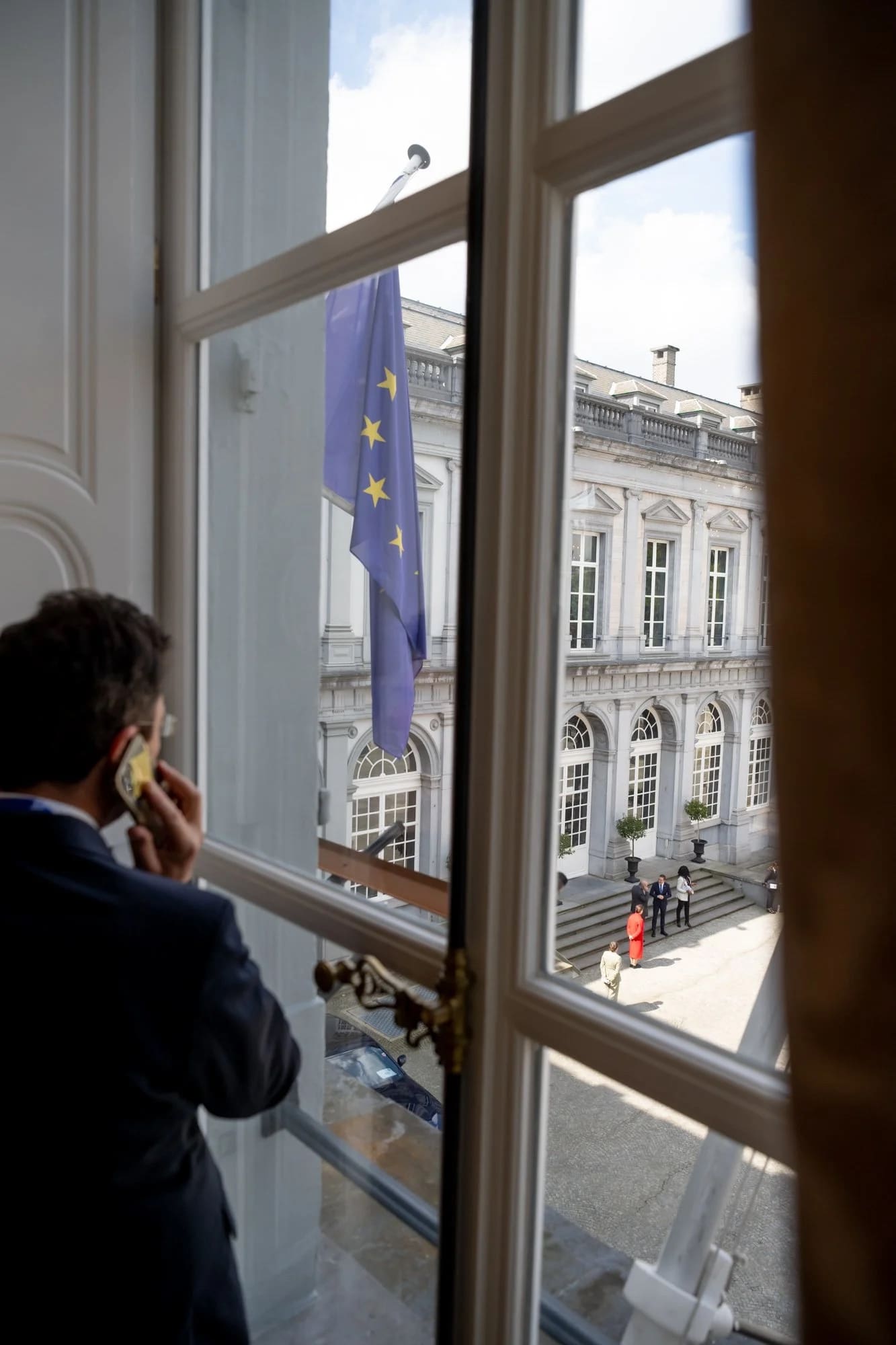 Een afgevaardigde telefoneert aan een hoog raam met zicht op de erekoer van een paleis.