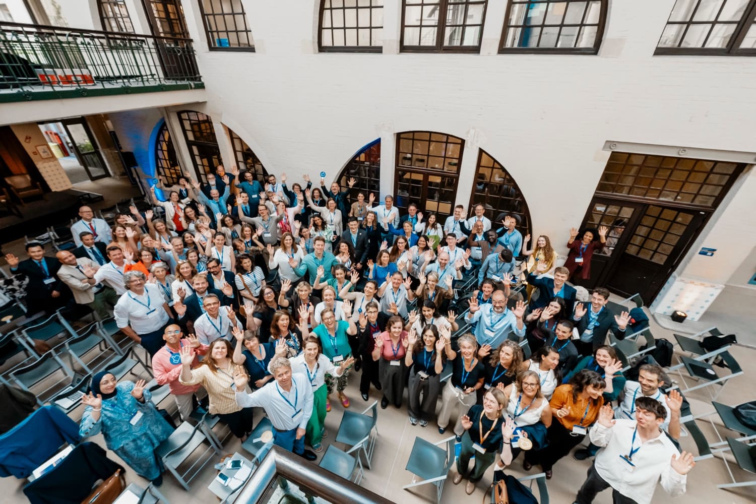 Overhead group photo: around a hundred participants wave at the camera in a courtyard.