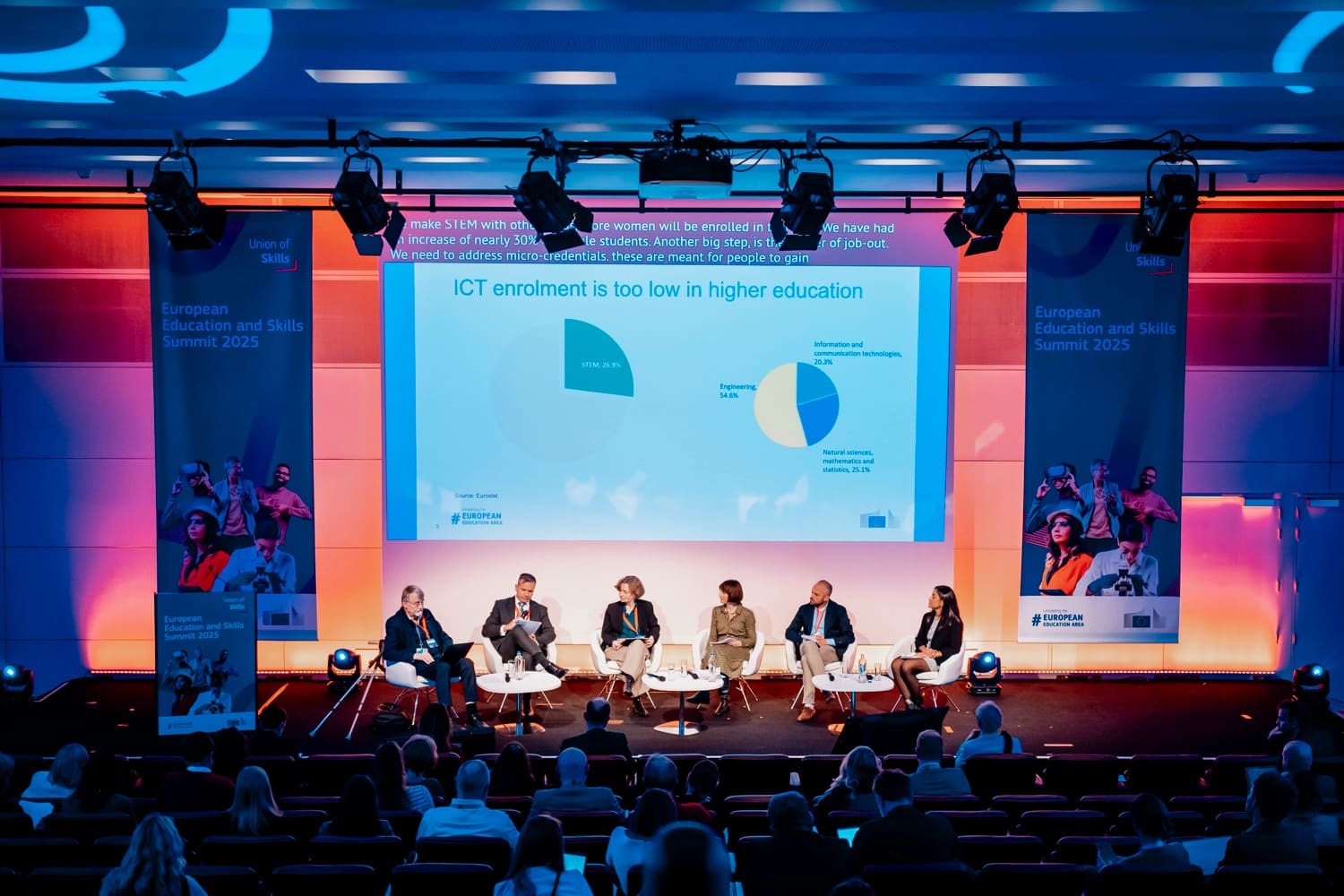Wide shot of a plenary: six panellists under a lighting rig, giant screen and audience.