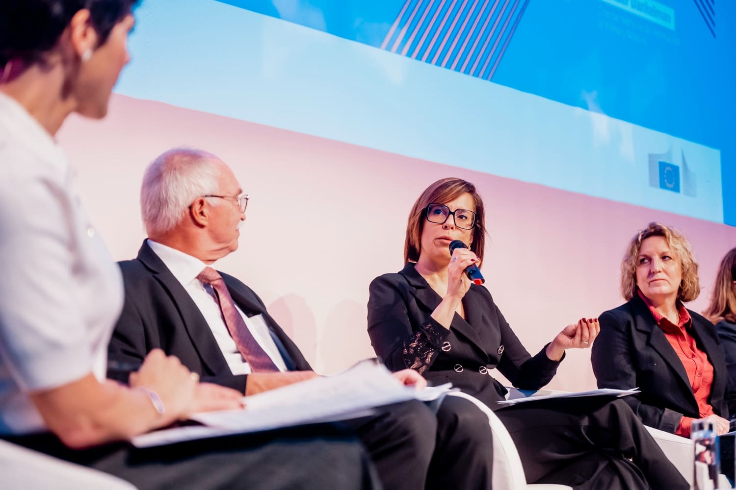 Panel on stage: a speaker at the microphone, her neighbours listening, pastel pink and blue backdrop.