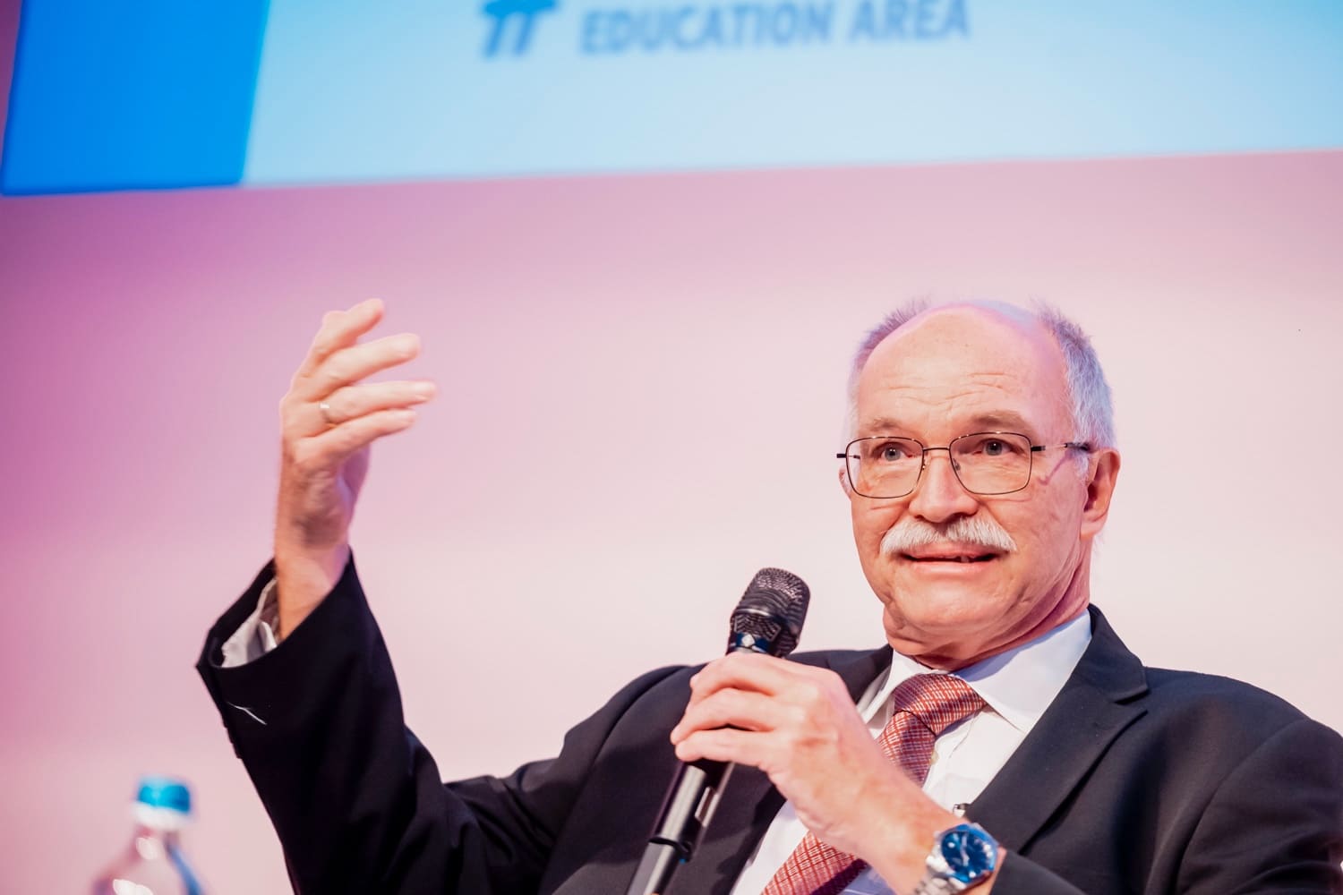 Panellist in glasses, microphone in hand, sweeping gesture, against a pastel pink backdrop.