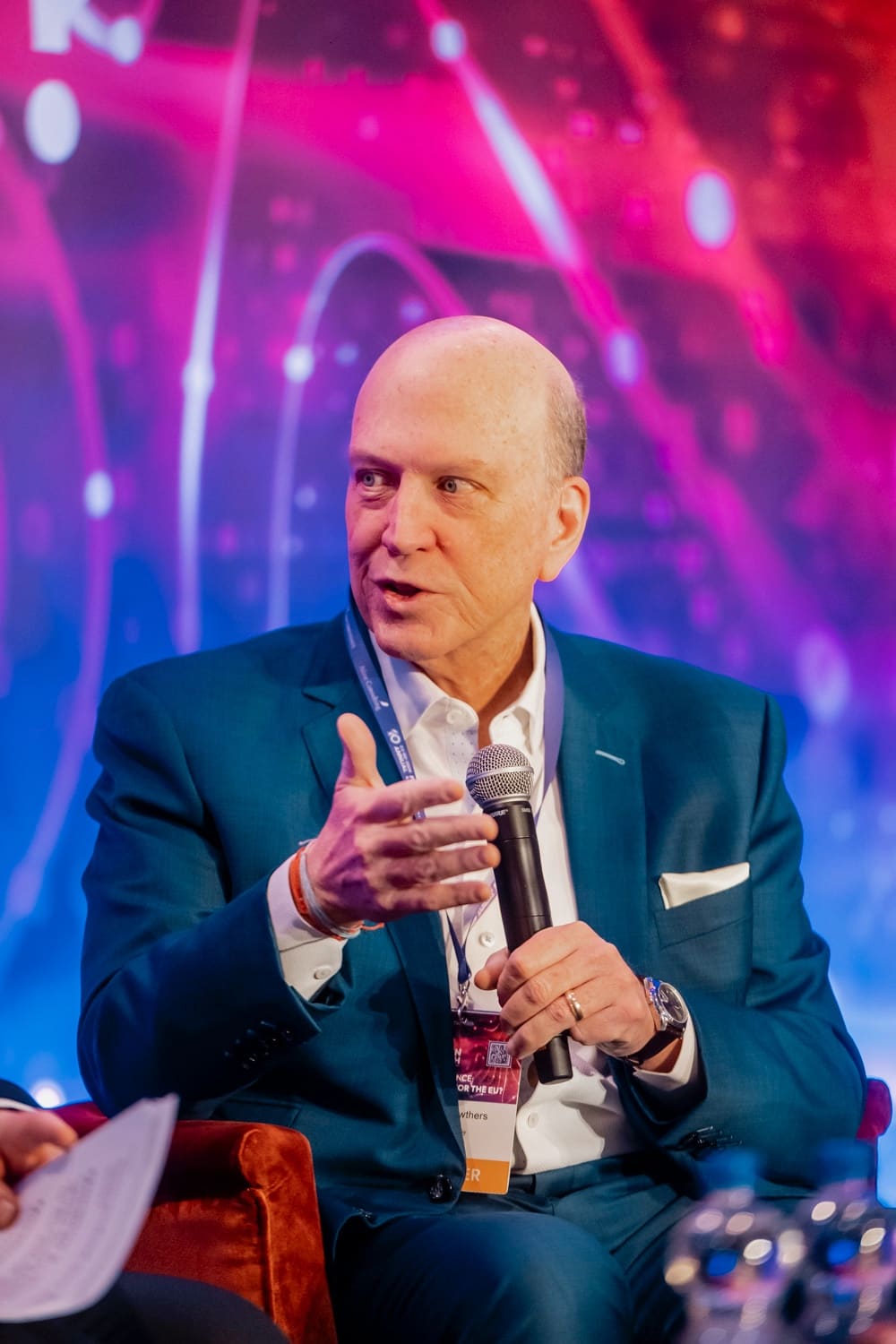 Panellist mid-explanation, microphone in hand, against a magenta and blue LED wall.