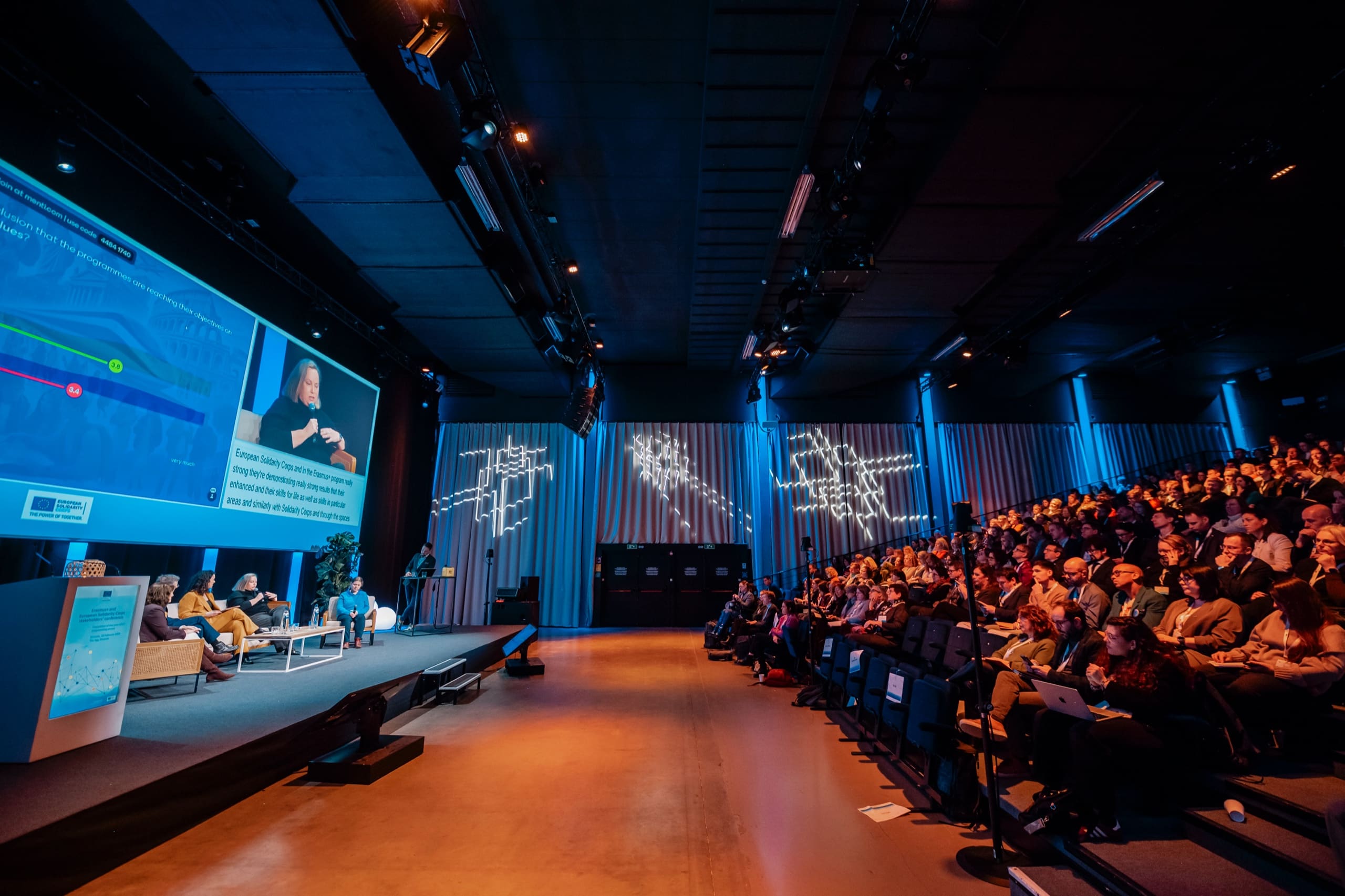 Auditorium en grand angle : panel sur canapés et écran géant à gauche, gradins pleins à droite.