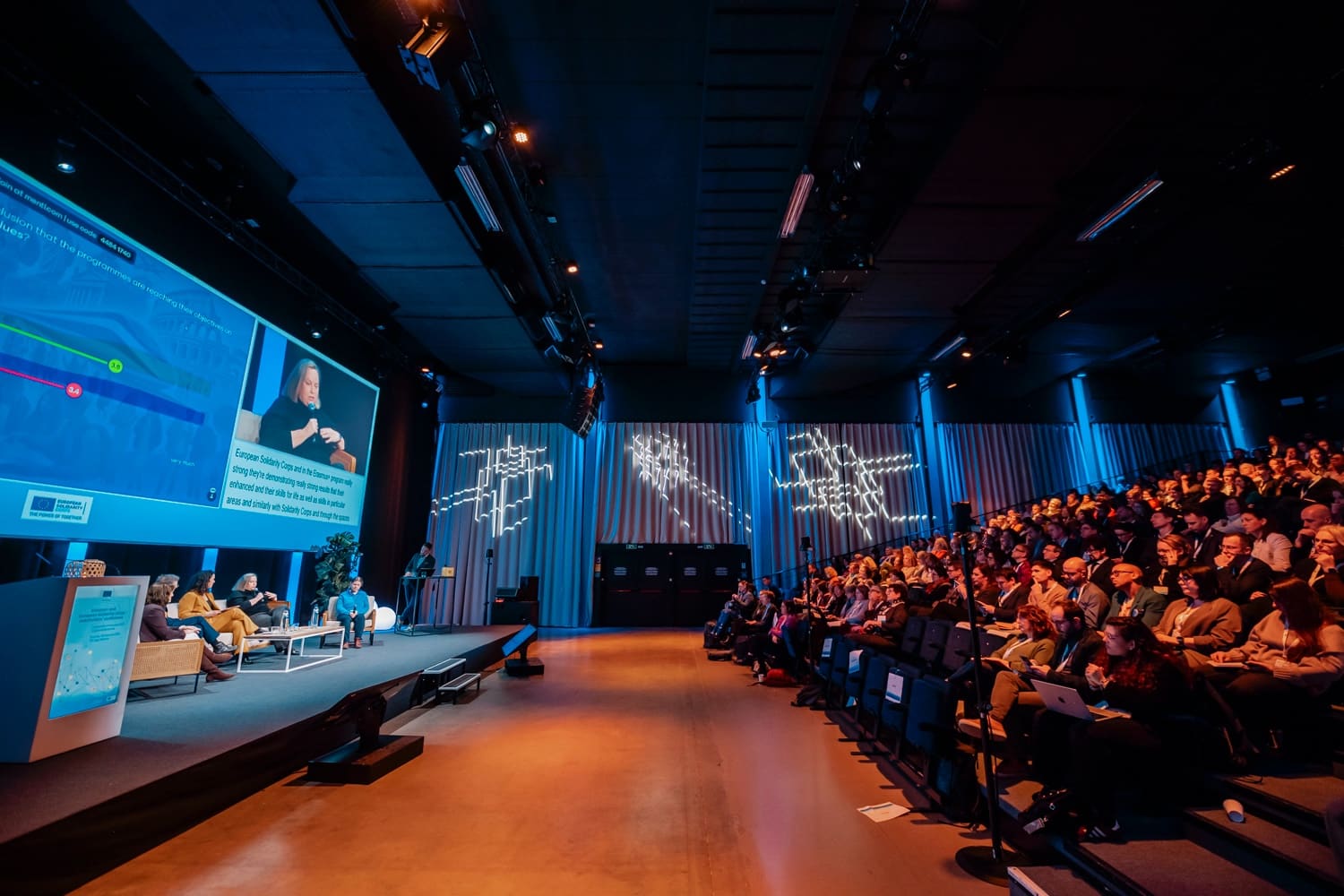 Wide-angle view of an auditorium: panel on sofas and giant screen on the left, packed tiers on the right.
