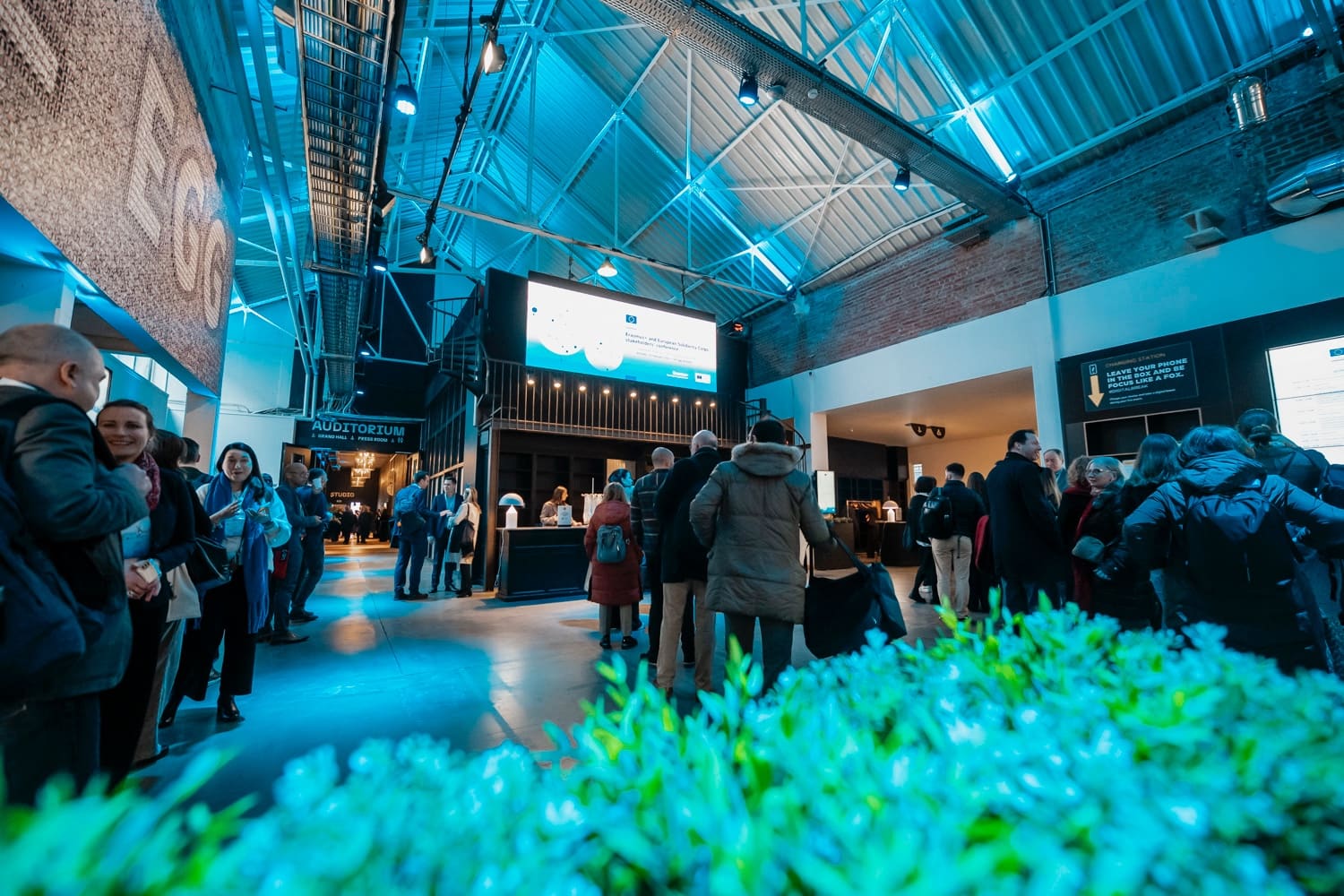 Industrial foyer full of participants, steel roof frame lit in cyan, glowing foliage in the foreground.