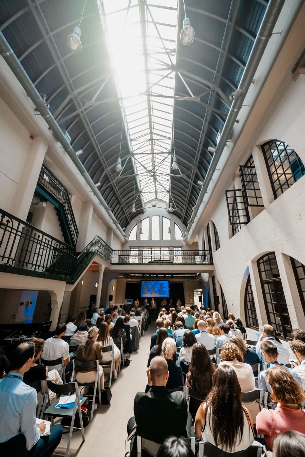 Former industrial hall with a glass roof: audience seated in rows towards the stage, wrought-iron galleries.