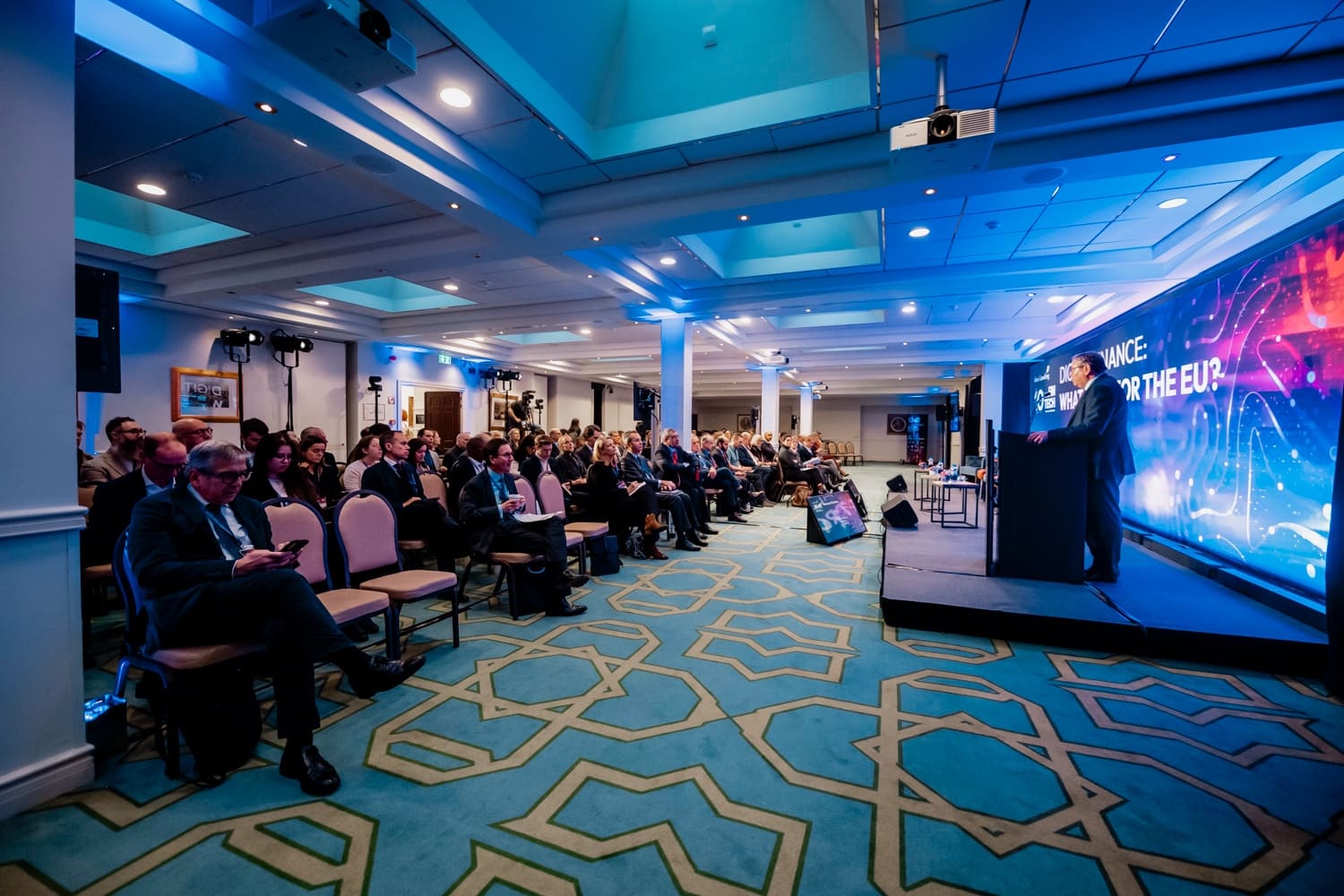 Wide-angle view of a conference room: seated audience, speaker on the platform facing the LED screen.