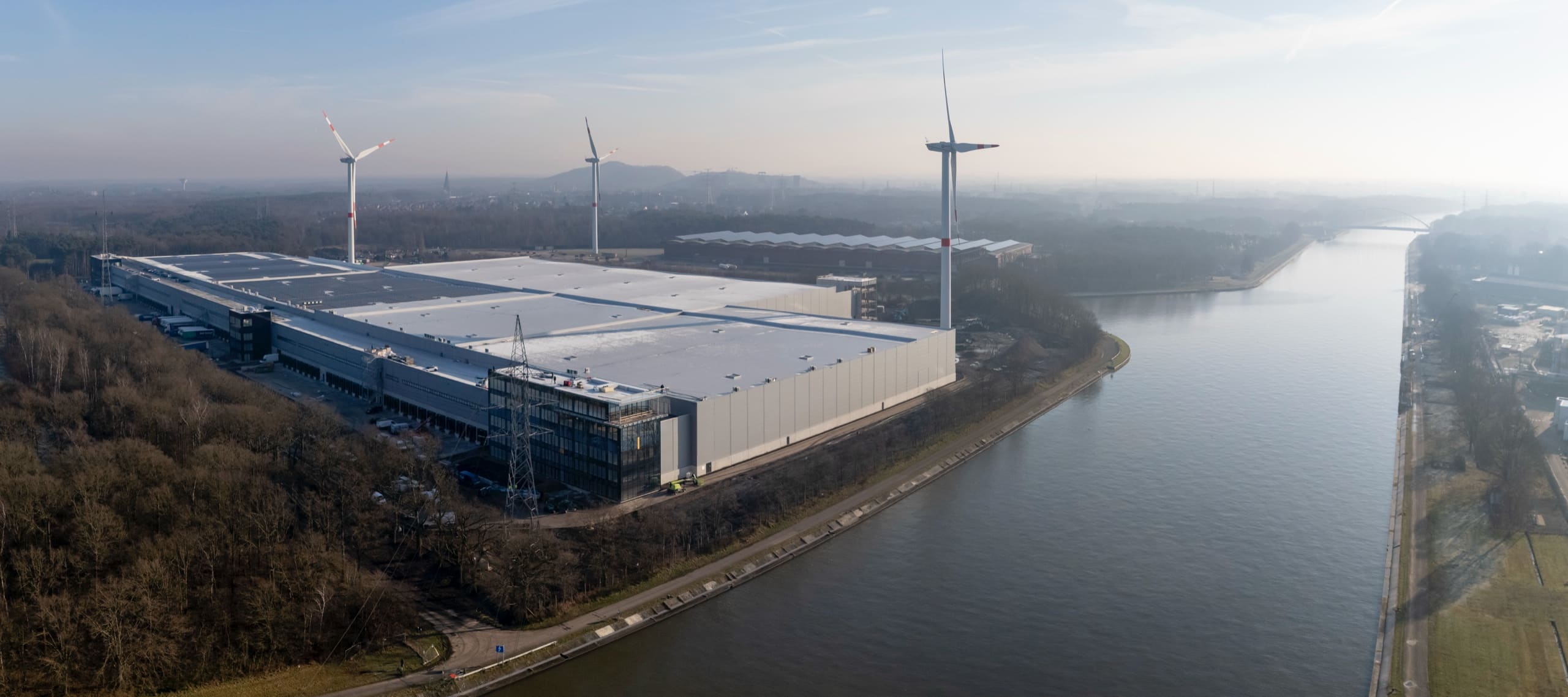 Panoramique aérien d’une plateforme logistique le long d’un canal, éoliennes et brume jusqu’à l’horizon.