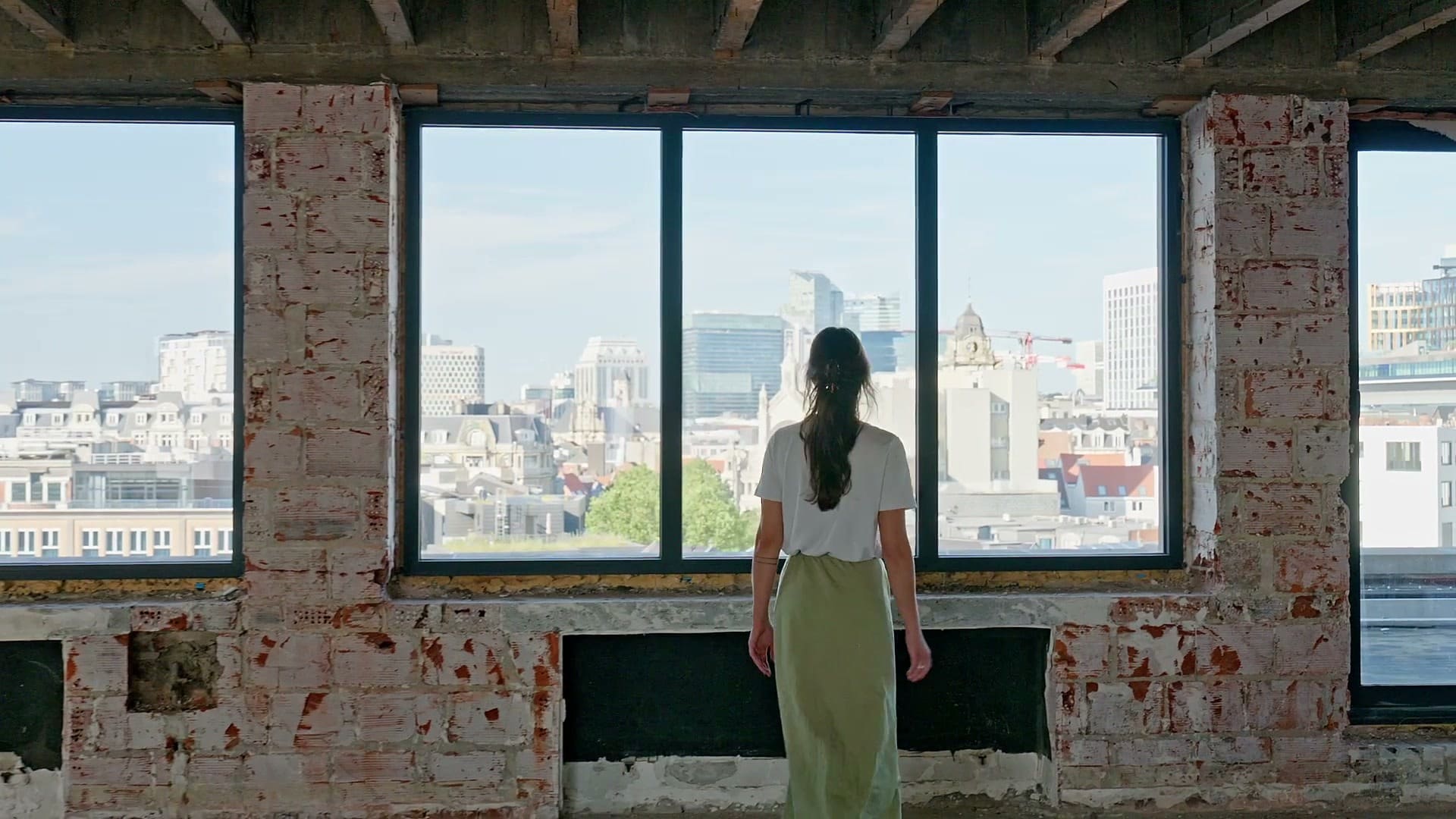 De dos, une femme regarde la ville par les grandes baies d’un étage en chantier.