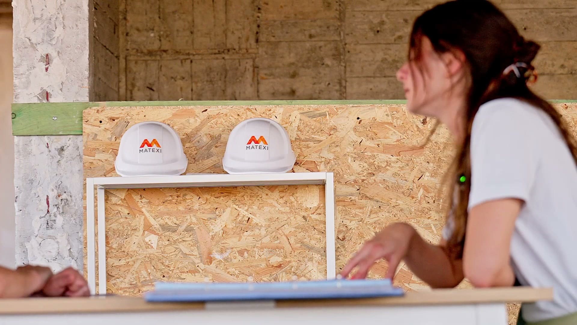 Des casques de chantier posés sur une étagère, une femme consulte un plan à côté.