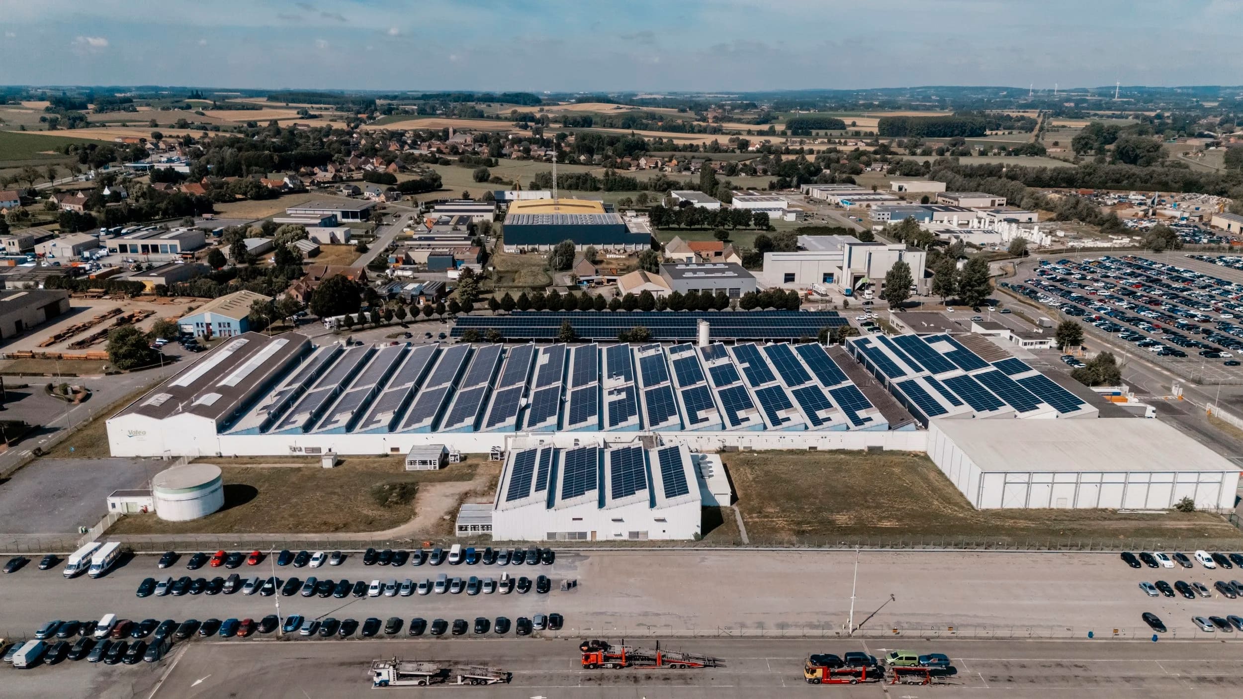 Luchtbeeld van een industriële site met zonnepanelen, omringd door parkings en platteland.