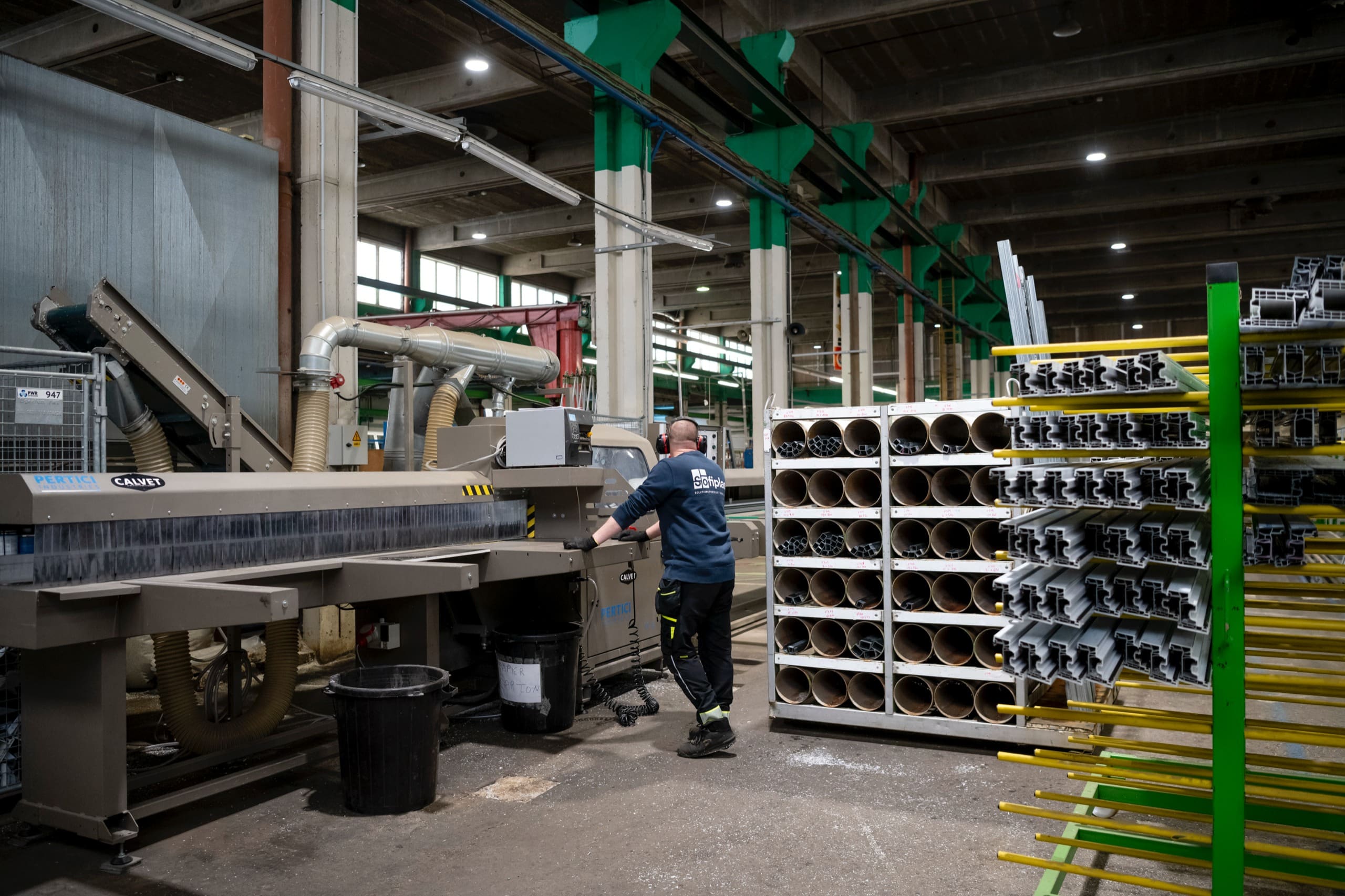 Le hall de production : une machine de découpe, des racks de profilés et un ouvrier au travail.
