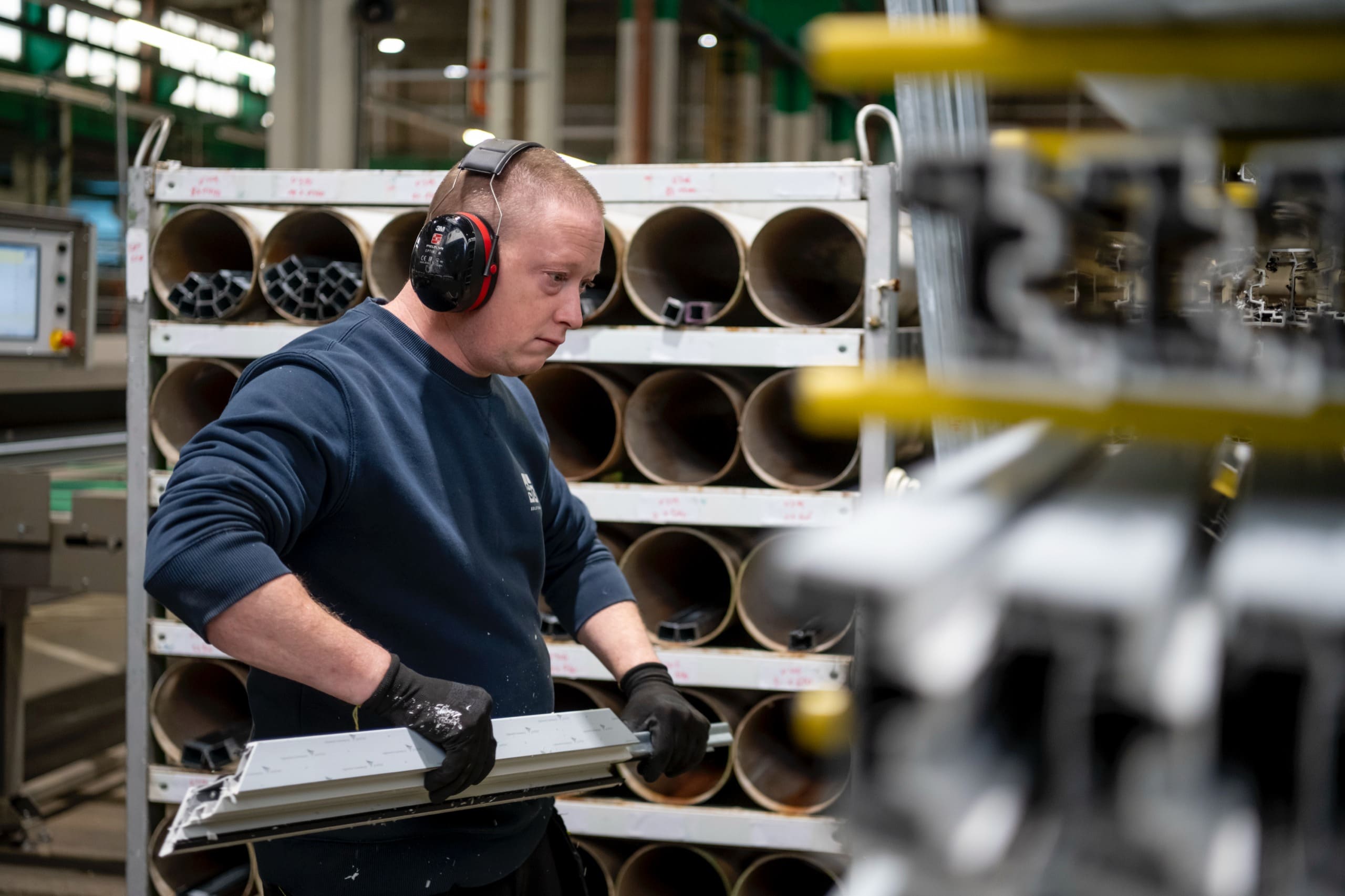 Un ouvrier en casque antibruit tient un profilé de châssis devant un rack de tubes.