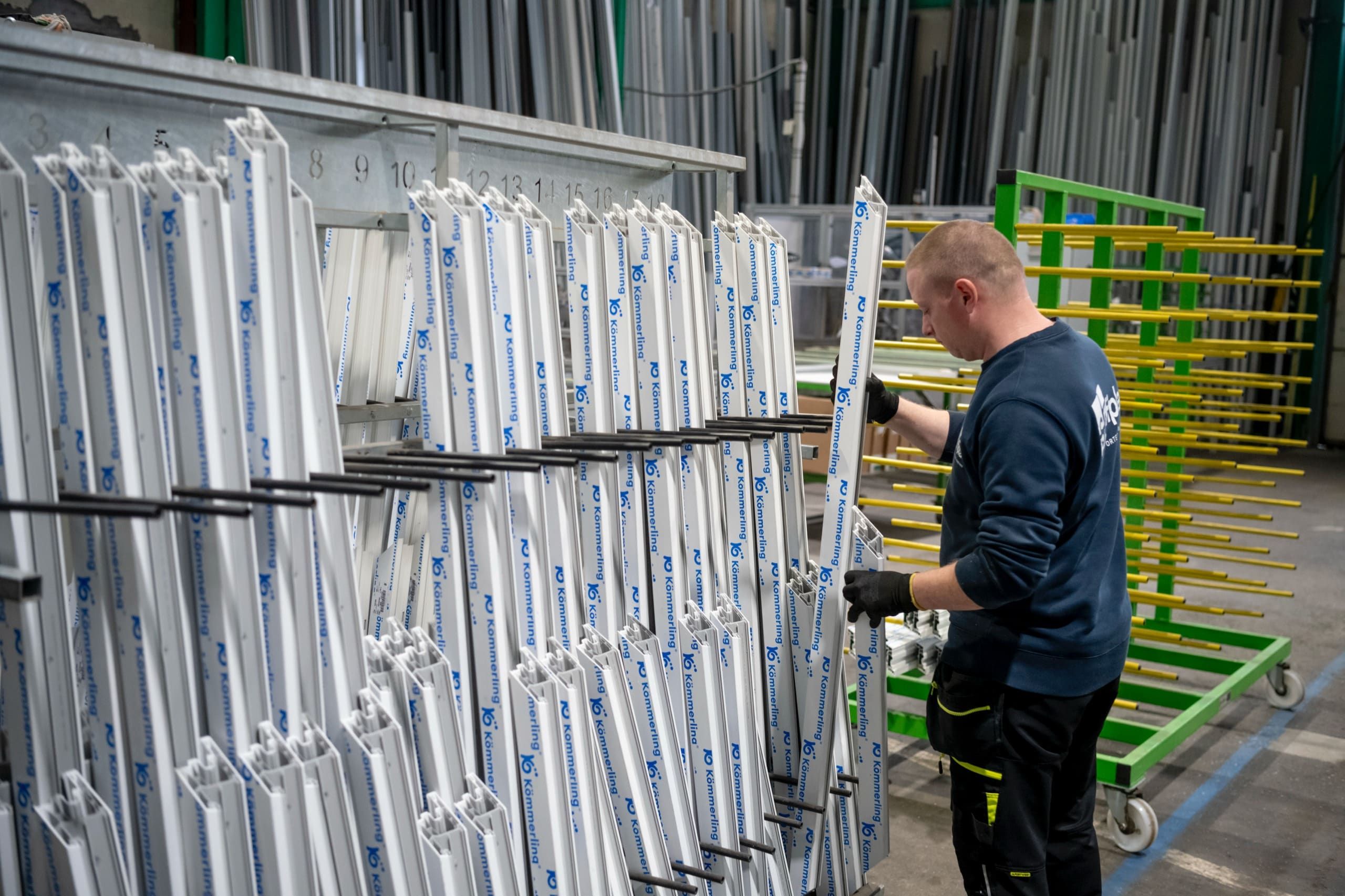 Un ouvrier range des profilés blancs dans un chariot, le long des racks verts.