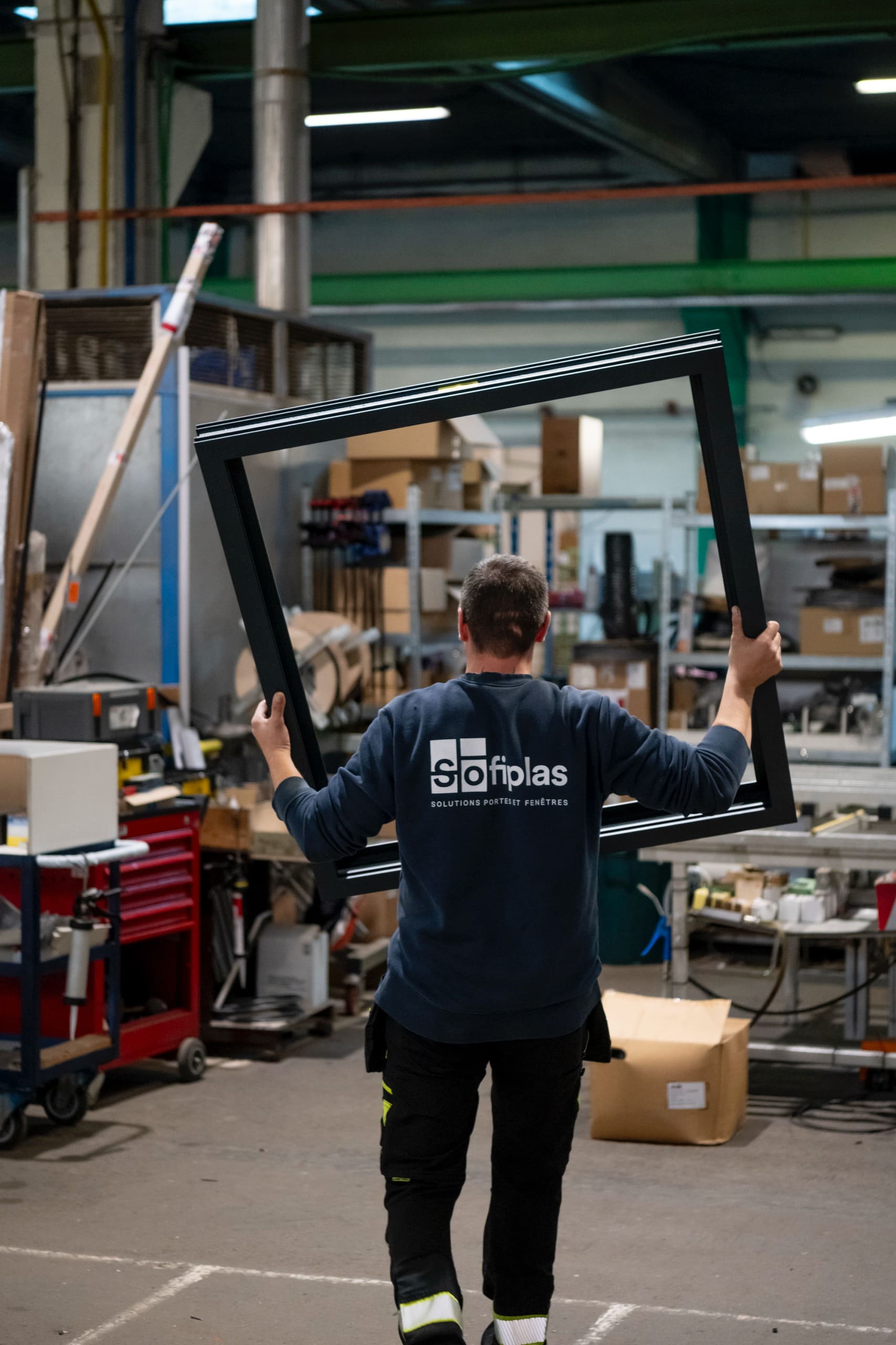 Un ouvrier porte un cadre de châssis noir à bout de bras dans l’atelier.