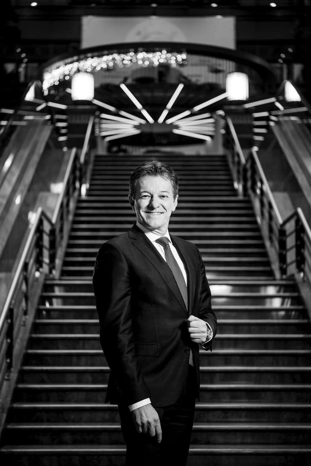 Black-and-white portrait of an executive at the foot of a grand staircase, beneath a radiant chandelier.