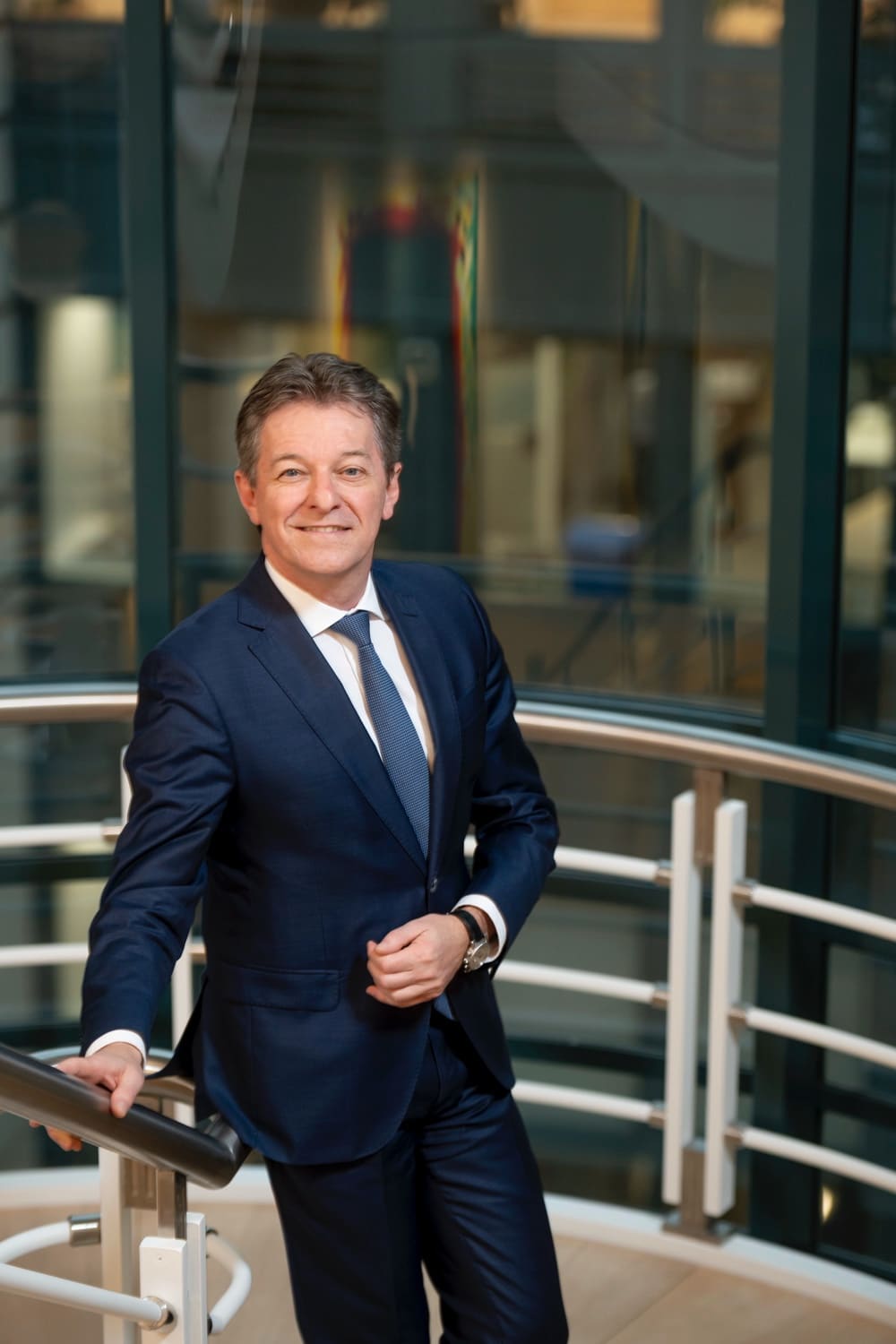 Portrait of an executive in a navy suit, leaning on a white railing in front of a glass atrium.