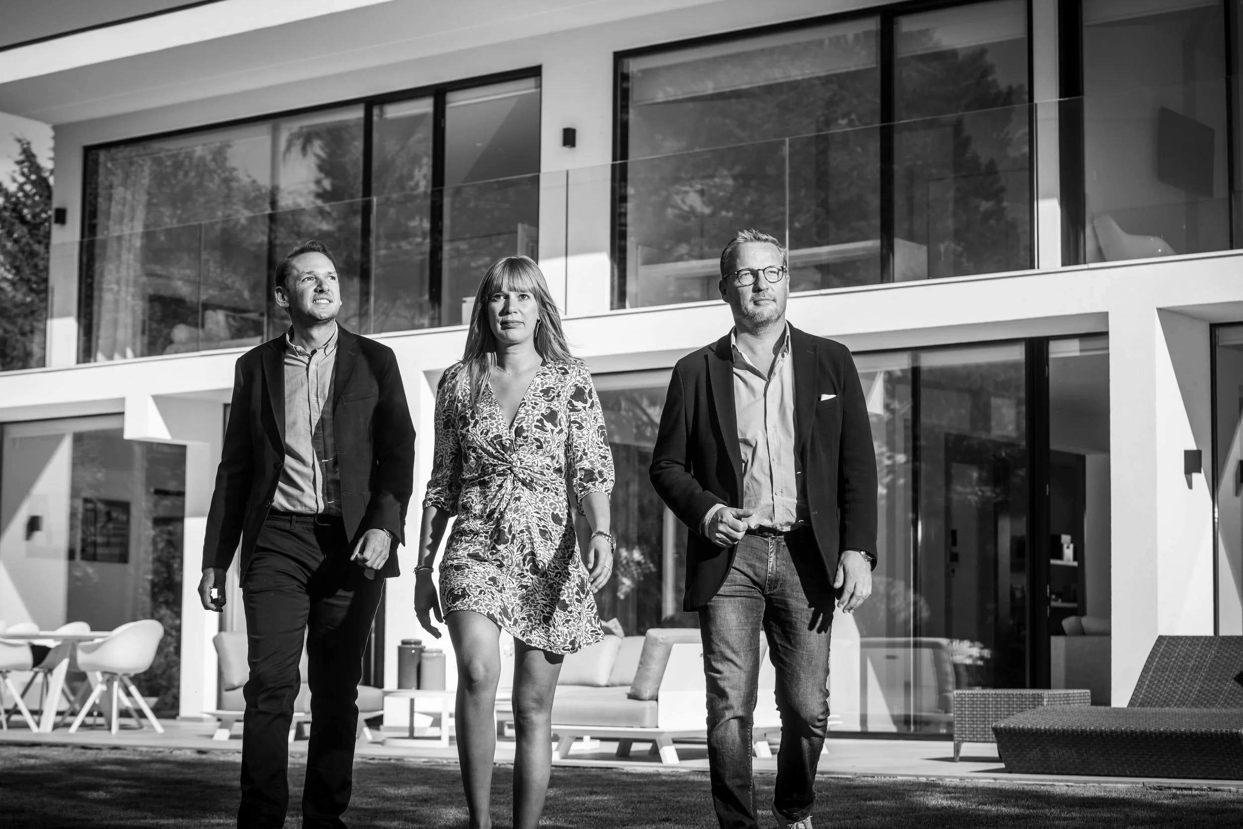 Black-and-white portrait of three partners walking towards the camera in front of a contemporary villa.